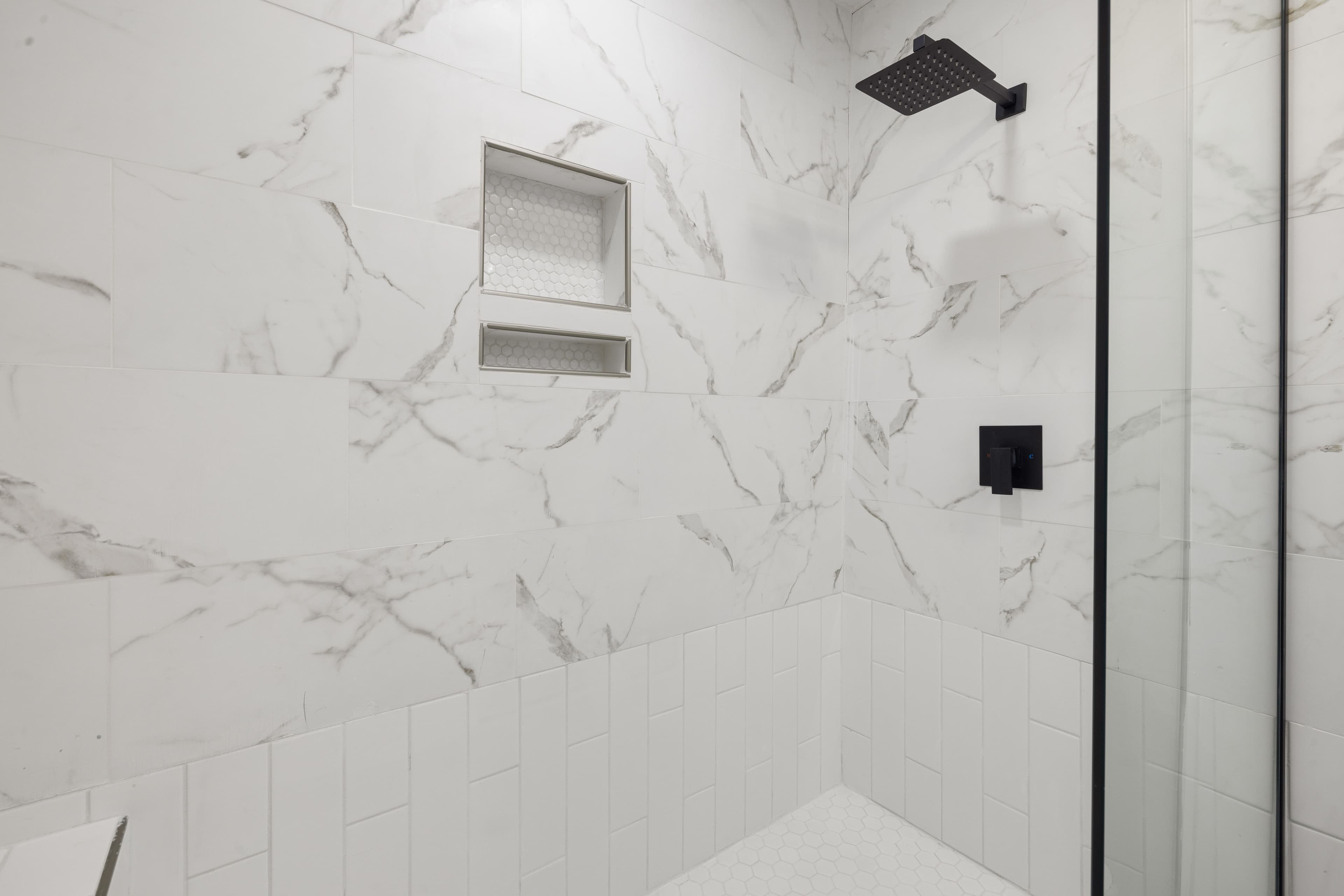 Walk-in shower design for a bathtub to shower conversion, with marble-look tile, recessed shelves and black fixtures.